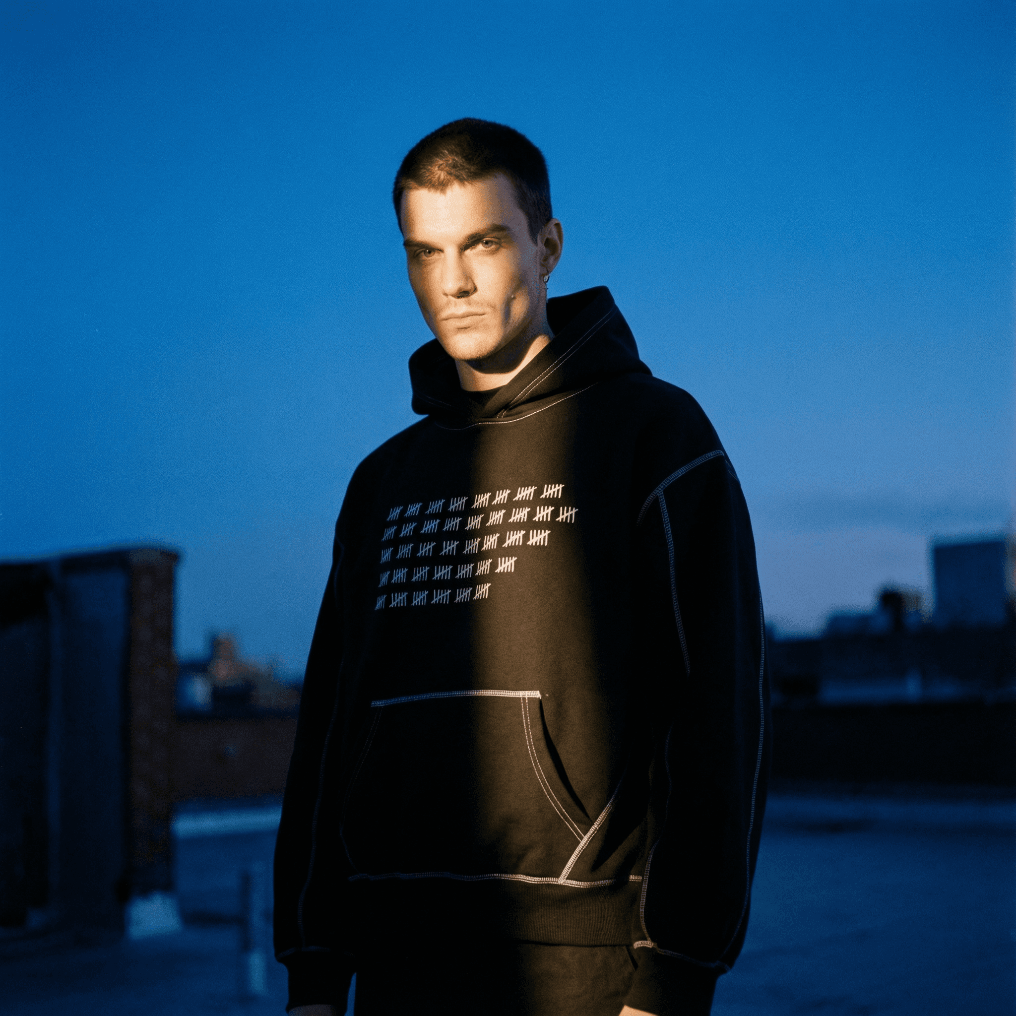 Man wearing black heavyweight fleece fetish hoodie with white stitch detailing and front pocket outdoors at dusk